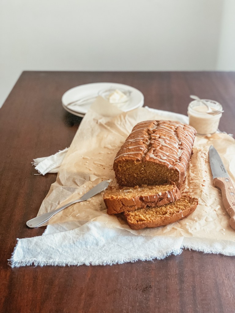 Pumpkin bread with maple icing sitting on parchment paper and a white cloth with butter and glaze in the background