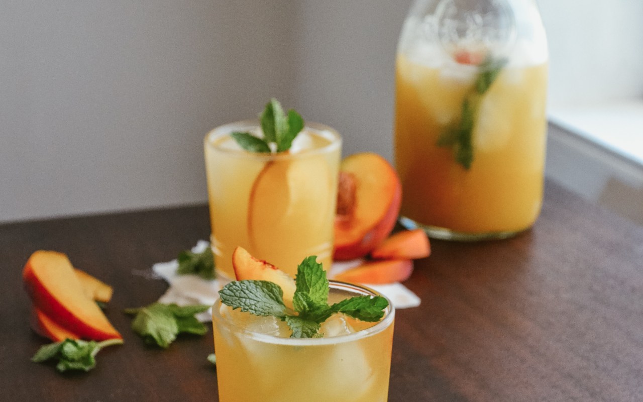 Peach mint white wine spritzer in a glass surrounded by peach slides and mint leaves with a pitcher full in the back