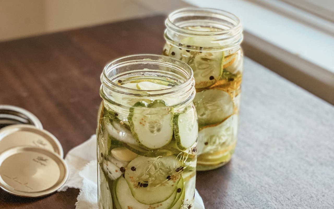 Two large jars of pickles on a wood surface in front of a white window