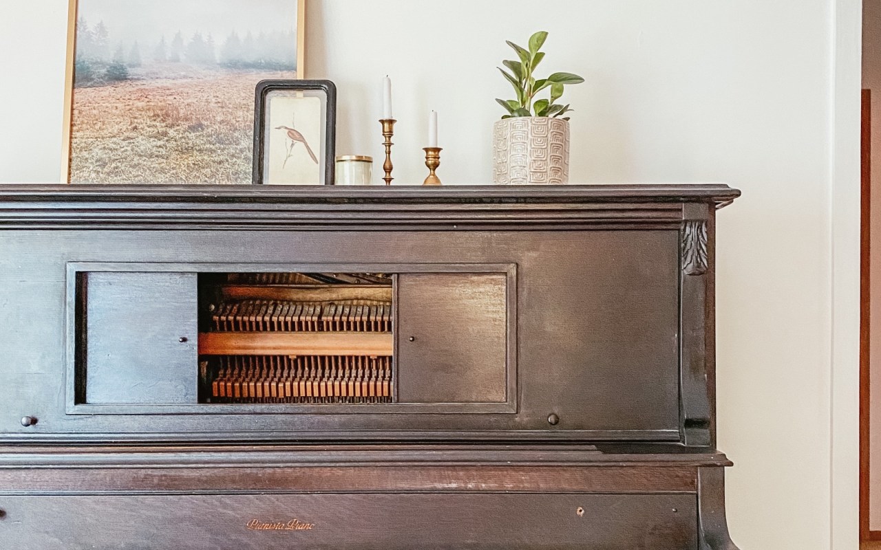 Dark brown stand up vintage piano with a plant, brass candle holders with pillar candles, a small framed drawing of a bird leaning against a large frame photo of a field all on top of the piano