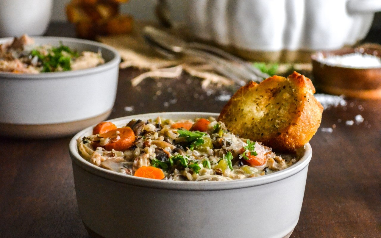 A bowl of creamy chicken with wild rice soup and a slice of parmesan herb toast another bowl sits behind and a pumpkin soup tureen