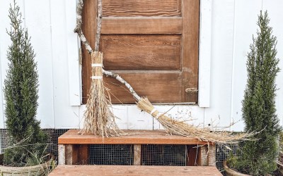 White chicken coop with two witch's brooms leaning against the door and pumpkins on the stairs
